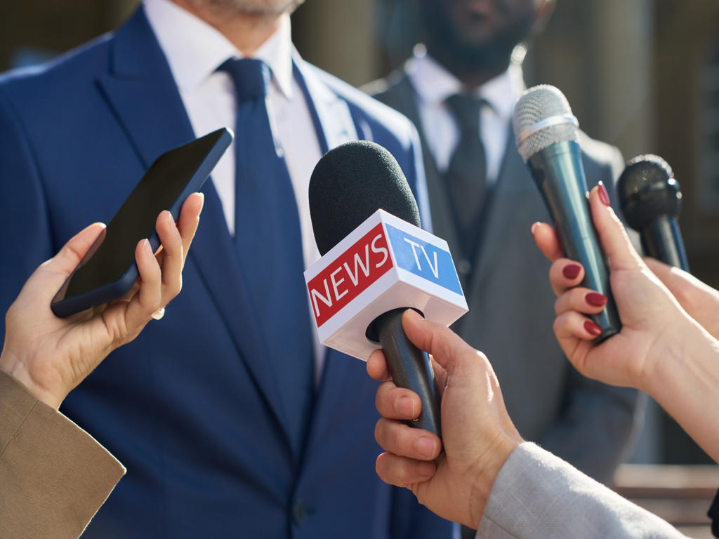 Caucasian middle aged man in suit speaking to group of journalists holding microphones and smartphone during outdoor press conference, hands of reporters visible in foreground